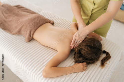 Closeup top view of pretty relaxed girl having neck, shoulder and back massage by unrecognizable female masseuse lying on massage table. Adorable kid getting physiotherapy from masseuse for body care