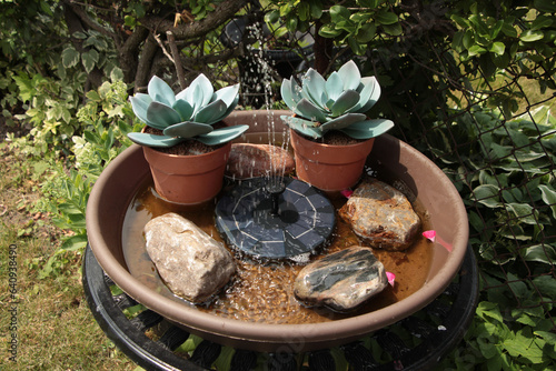 small black solar power fountain spraying water up in brown circular bin with rocks and fake plants in it on top of a table with grass and greenery in the background