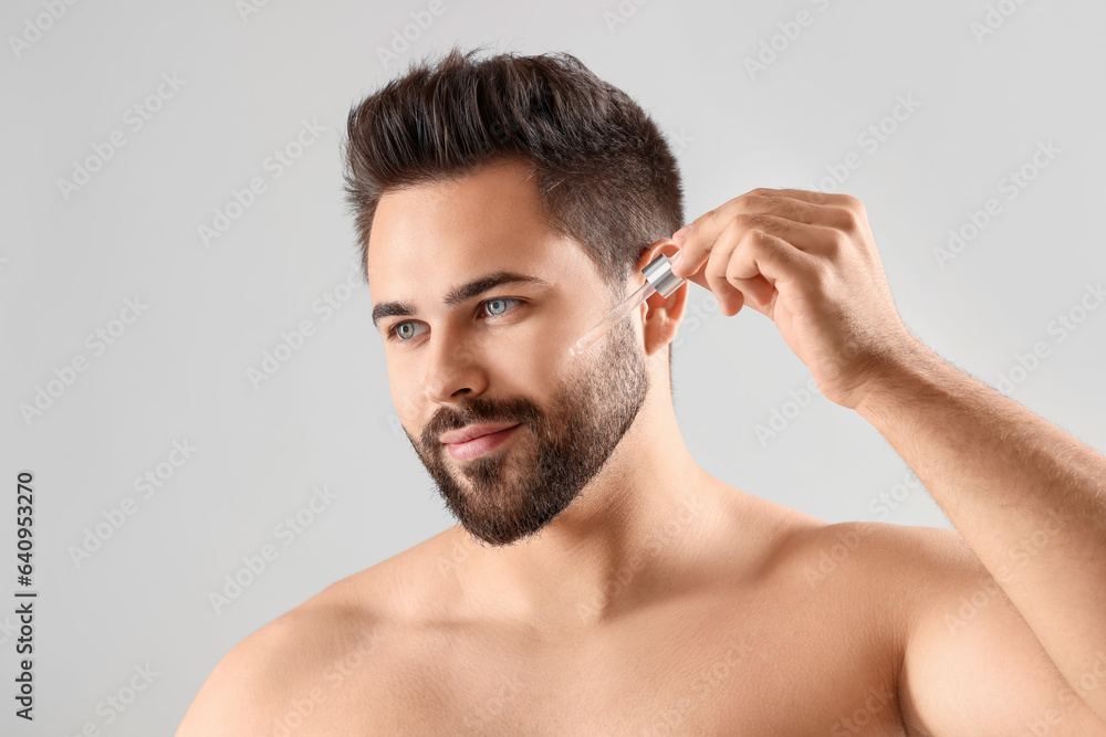 Obraz premium Handsome man applying cosmetic serum onto face on light grey background