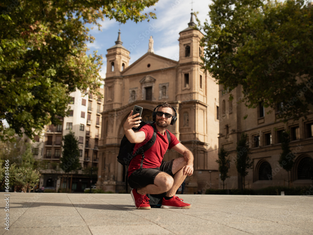 A young male traveler and backpacker, wearing sunglasses, smiles at the camera, while taking a selfie with his cell phone during a sightseeing tour of a Spanish city.