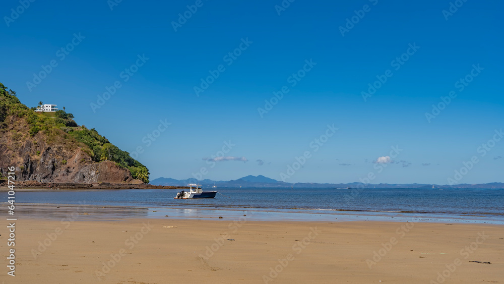 Sandy beach on a tropical island. The boat is moored in the   blue ocean. The white building on the hill. A mountain range against the azure sky on the horizon. Madagascar. Nosy Be island.
