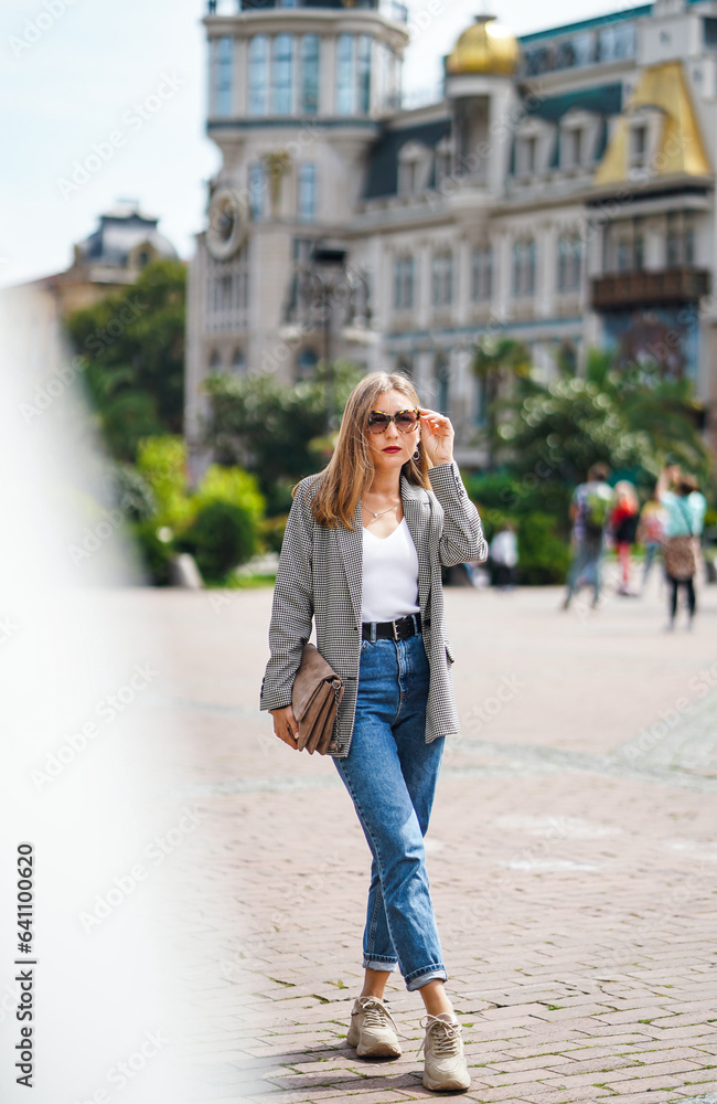Bright sunny day. Girl at fountain. Businesswoman wearing jacket and jeans holds bag in square of Batumi city in Georgia. Behind is old beautiful architectura building. Girl walks around city