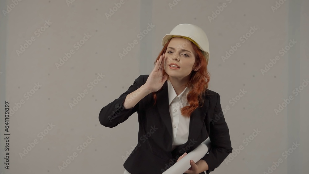 A woman architect, putting her hand to her mouth, screams, someone's advice. Portrait of a woman in a protective helmet and with blueprints in the studio on a gray background. A woman architect, putting her hand to her mouth, screams, someone's advice. Portrait of a woman in a protective helmet and with blueprints in the studio on a gray background.