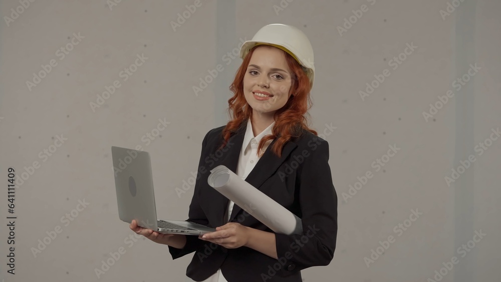 A woman architect works on a laptop, studies plans, prints reports. Business woman in a protective helmet, with drawings under her arm and a laptop on a gray background in the studio. A woman architect works on a laptop, studies plans, prints reports. Business woman in a protective helmet, with drawings under her arm and a laptop on a gray background in the studio.