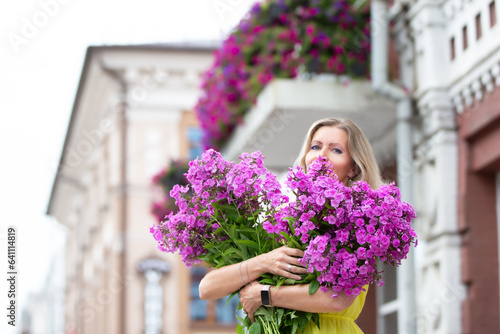 A beautiful elderly woman with a bouquet of flowers walks around the city. Age model blond with blue eyes is happy. A woman of fifty years walks in the summer, having a good time.