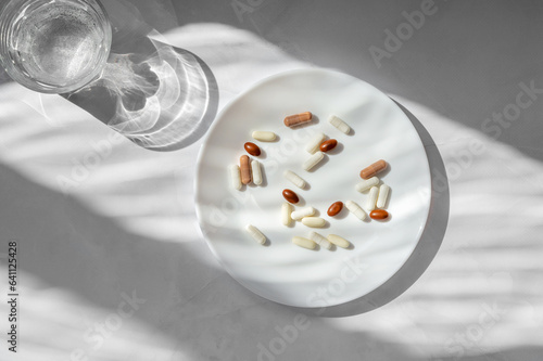 Vitamin and supplement pills and capsules on white plate, glass with pure water on marble table with natural sunlight shadows. Healthcare, beauty and pharmaseutical industry lifestyle concept