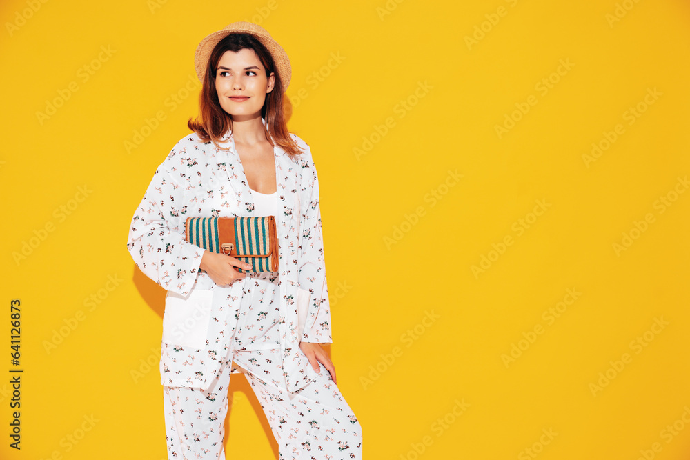 Obraz premium Young beautiful smiling female in trendy summer costume clothes. Carefree woman posing near yellow wall in studio. Positive model having fun indoors. Cheerful and happy. In hat. Holds clutch