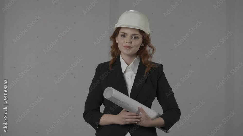 Portrait of a young woman architect with blueprints in her hand close up. Woman architect in a protective helmet and strict suit in the studio on a gray background. Architectural project. HDR BT2020 Portrait of a young woman architect with blueprints in her hand close up. Woman architect in a protective helmet and strict suit in the studio on a gray background. Architectural project. HDR BT2020
