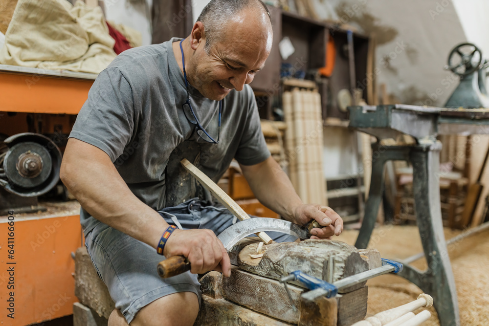 Happy adult ethnic man carpenter sitting and using sickle to carve wood in workshop