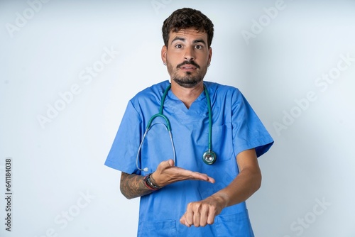 young caucasian doctor man wearing blue medical uniform In hurry pointing to watch time, impatience, upset and angry for deadline delay.