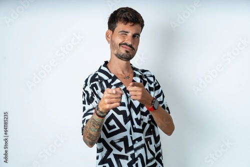 Canvas Print Joyful young caucasian man wearing printed shirt wink and points index fingers at camera, chooses someone, makes finger gun pistol