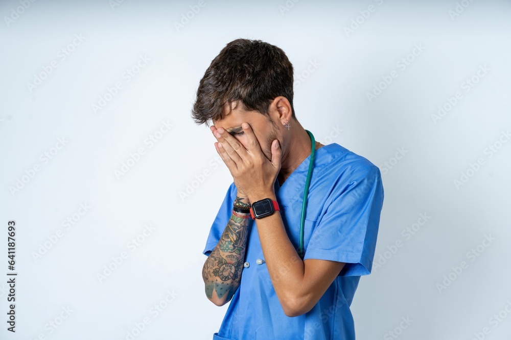 Sad young caucasian doctor man wearing blue medical uniform crying covering her face with her hands. Sad young caucasian doctor man wearing blue medical uniform crying covering her face with her hands.