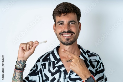 young caucasian man wearing printed shirt holding an invisible aligner and pointing at it. Dental healthcare and confidence concept.