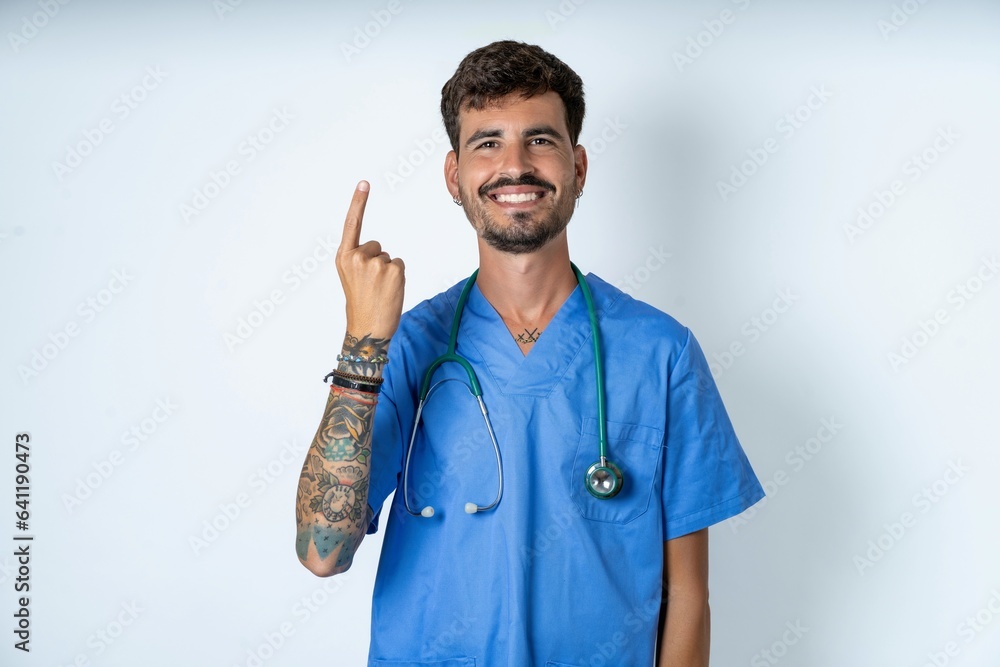 young caucasian doctor man wearing blue medical uniform smiling and looking friendly, showing number one or first with hand forward, counting down young caucasian doctor man wearing blue medical uniform smiling and looking friendly, showing number one or first with hand forward, counting down