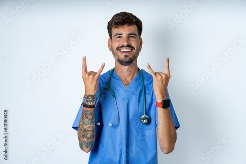 young caucasian doctor man wearing blue medical uniform makes rock n roll sign looks self confident and cheerful enjoys cool music at party. Body language concept.