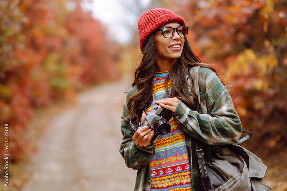 Happy female tourist with a camera and a backpack in a stylish sweater on a walk in the autumn forest. A woman takes pictures and enjoys the beautiful nature. Happy female tourist with a camera and a backpack in a stylish sweater on a walk in the autumn forest. A woman takes pictures and enjoys the beautiful nature.