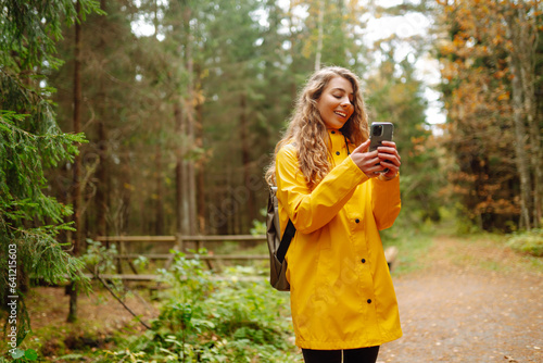 Young woman traveler in a yellow raincoat, holding a phone in her hands against the backdrop of a forest thicket. Smiling woman uses a mobile phone while walking in the forest.