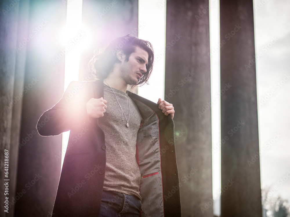 Photo of a man standing in front of tall pillars holding a jacket Photo of a man standing in front of tall pillars holding a jacket