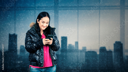 Portrait of a happy young teenage girl dressed in fur jacket using mobile phone over snowy city