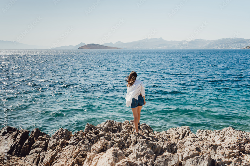 Woman in denim shorts and a white shirt, stands on the shore of the blue sea on the rocks.