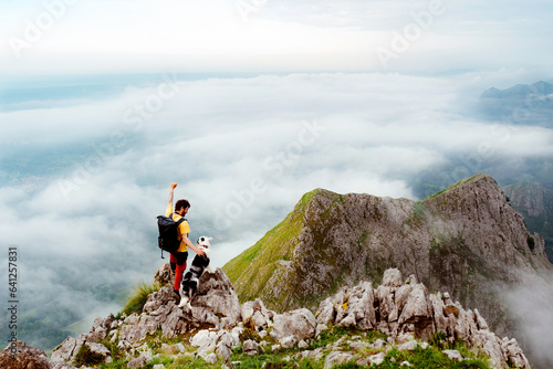 white male mountaineer standing on a rock next to his dog contemplating the mountain landscape covered by a sea of clouds after a day of hiking.