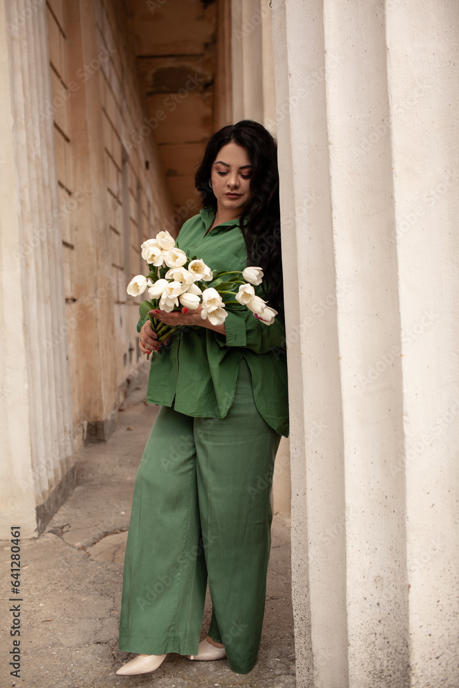 Fototapeta premium Beautiful woman holding the bouquete of white flowers. The plus size woman dresssed urban style outfit, green pajamas style suit. The overweiht woman with long hairs