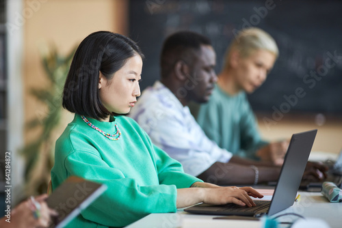 Young serious Asian female student in green pullover looking at laptop screen and typing while analyzing or decoding data against classmates