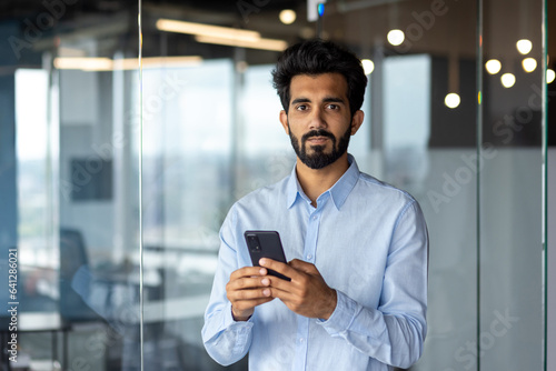 Portrait of young Indian male manager, office worker standing indoors and using phone. He looks seriously into the camera