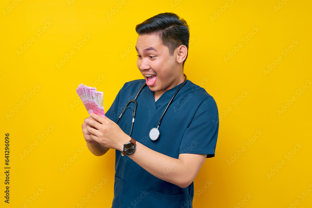 Portrait of a smiling excited young Asian male doctor or nurse wearing blue uniform holding cash money isolated on yellow background. Healthcare medicine concept Portrait of a smiling excited young Asian male doctor or nurse wearing blue uniform holding cash money isolated on yellow background. Healthcare medicine concept