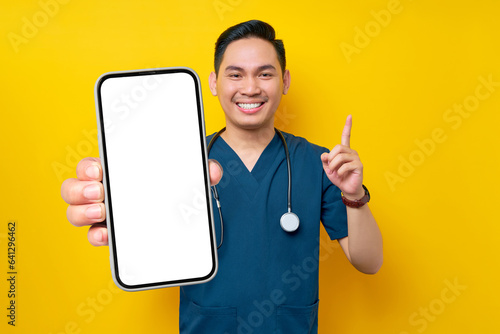 Smiling young Asian male doctor or nurse wearing blue uniform showing a smartphone blank screen and pointing a finger up isolated on yellow background. Healthcare medicine concept