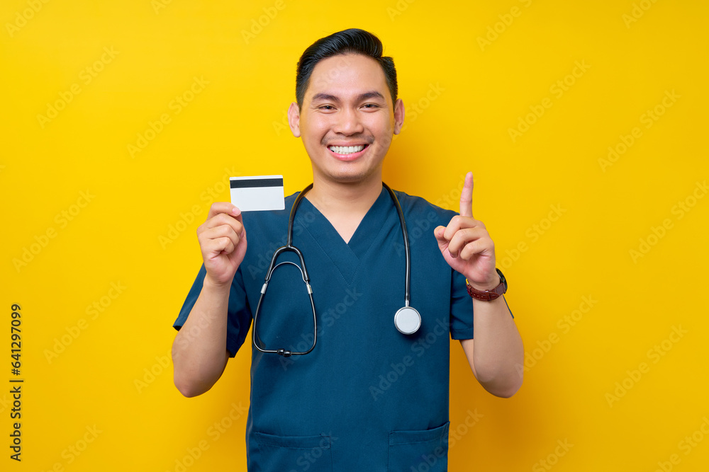 Smiling cheerful young Asian male doctor or nurse wearing blue uniform holding credit bank card and pointing finger up copy space isolated on yellow background. Healthcare medicine concept Smiling cheerful young Asian male doctor or nurse wearing blue uniform holding credit bank card and pointing finger up copy space isolated on yellow background. Healthcare medicine concept