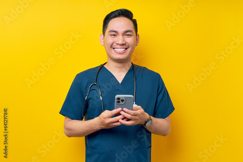 Smiling happy young Asian male doctor or nurse wearing blue uniform holding mobile phone and looking at a camera isolated on yellow background. Healthcare medicine concept