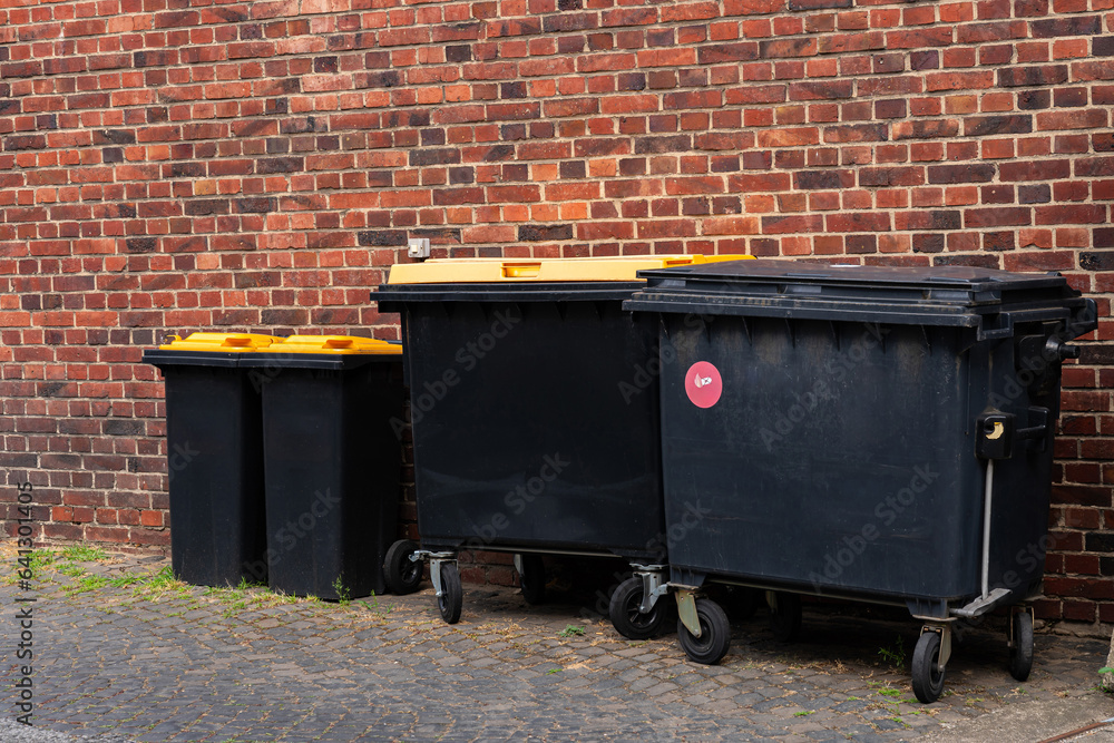 Black plastic garbage cans with yellow lids standing near the wall of a brick building. Black plastic garbage cans with yellow lids standing near the wall of a brick building.