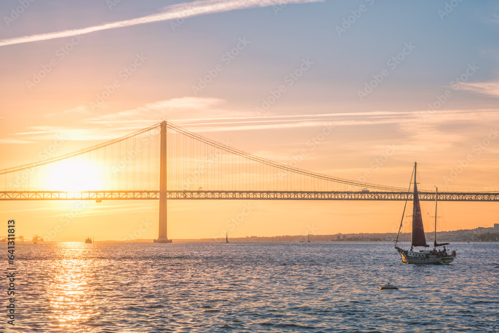 View of 25 de Abril Bridge famous tourist landmark of Lisbon connecting Lisboa and Almada over Tagus river with tourist yacht silhouette at sunset. Lisbon, Portugal View of 25 de Abril Bridge famous tourist landmark of Lisbon connecting Lisboa and Almada over Tagus river with tourist yacht silhouette at sunset. Lisbon, Portugal