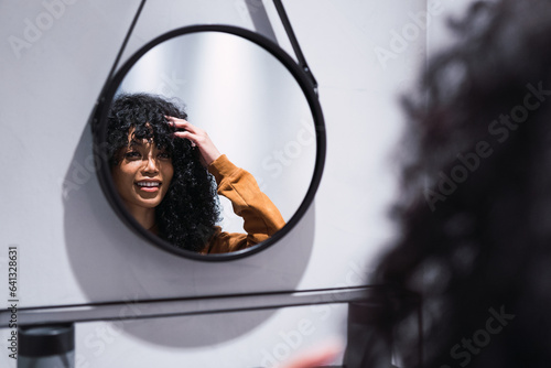 Young brunette woman adjusting hair and smiling while looking at mirror in bathroom.