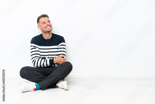 Young man sitting on the floor isolated on white background looking up while smiling
