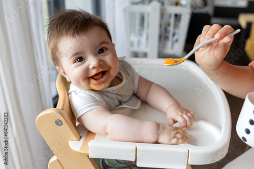 Cuadro en lienzo Happy baby boy eating first food pumpkin from spoon with mom at home