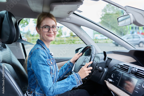 Teenage girl driver in glasses sitting behind wheel of car, looking at camera