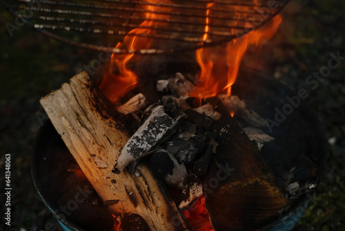 burning fire in compact grill, wood logs engulfed red flames, closeup of metal grill on burning coals, aromatic smoke rises appetizingly, fun party, happy childhood, family activity, cooking outdoors
