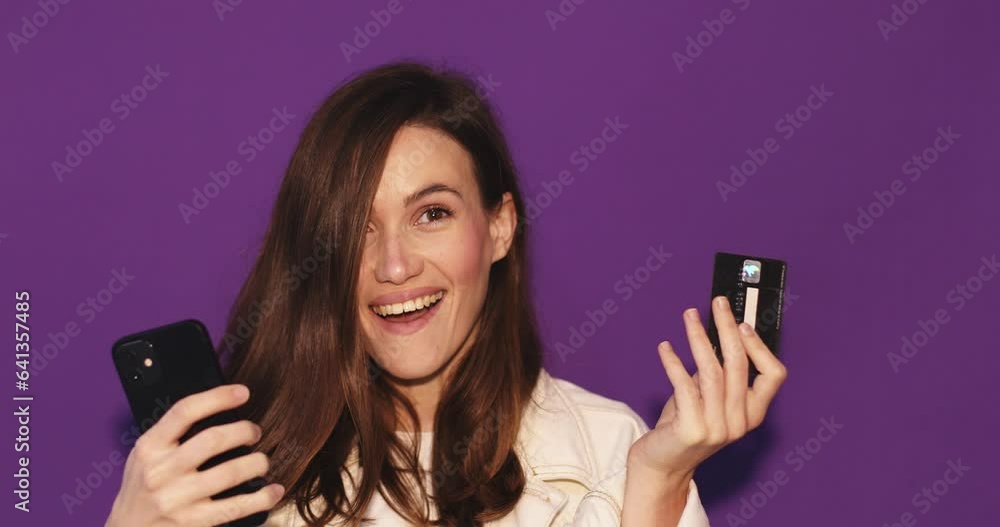Young woman wear white jeans jacket using mobile cell phone hold in hand credit bank card do online shopping book tour isolated on purple background. Wow surprised girl look at phone, show credit card Young woman wear white jeans jacket using mobile cell phone hold in hand credit bank card do online shopping book tour isolated on purple background. Wow surprised girl look at phone, show credit card