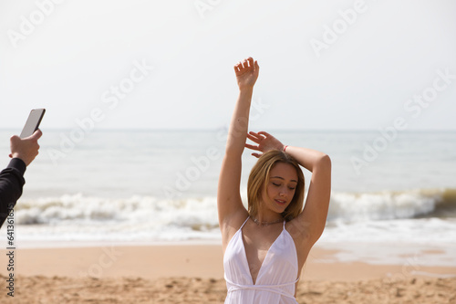 Young beautiful blonde woman in white dress is walking on the sand on the shore of the beach on a sunny day. The woman makes different body expressions. In the background the blue sea.