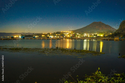 The evening atmosphere at Lake Batur, Kintamani, Bali, with the reflection of Mount Batur and the ambiance of the village along the lake's edge, illuminated by sparkling lights.