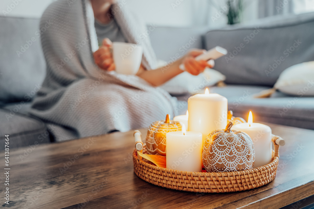 Focus on autumn fall cozy mood decor composition with a blurred woman covered plaid selecting movie with remote controller and drinking tea on the sofa. Movie night party at home. Cozy relaxing time.