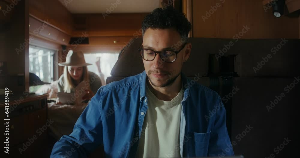 A young man uses a laptop and drinks a hot drink from a metal mug while sitting at a table in a van. A woman reads a book in the background A young man uses a laptop and drinks a hot drink from a metal mug while sitting at a table in a van. A woman reads a book in the background