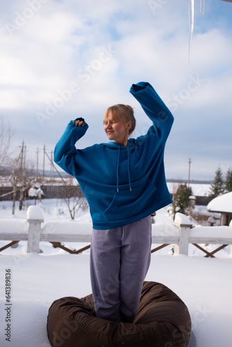 A young girl in a blue hoodie is sitting on a brown ottoman in the snow on the balcony in winter