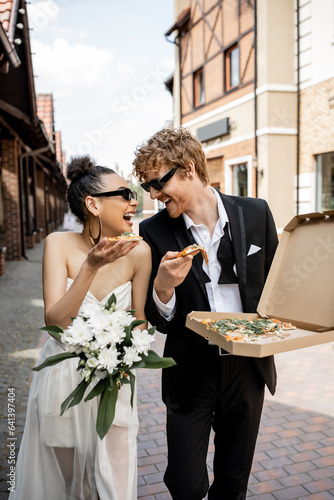 Fotografía young multiethnic newlyweds with pizza and flowers laughing on city street, outd