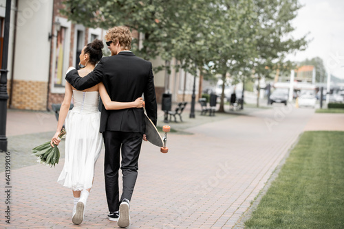 back view of multiethnic elegant newlyweds with bouquet and longboard embracing and walking in city