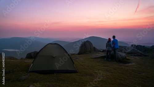 Couple watching red, purple sunset. Tent on the top of mountain. Panoramic view of the Mourne Mountains, beautiful part of Northern Ireland