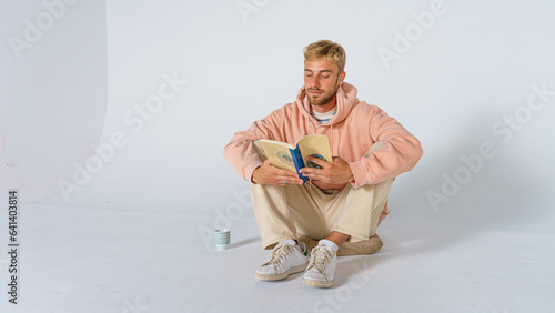 blond man in casual clothes on a white background sitting on the floor on a cushion reading a book and drinking a beverage