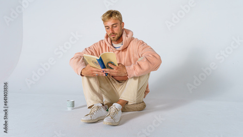 handsome concentrated blond man in casual clothes on a white background sitting on the floor on a cushion reading a book and drinking a beverage